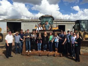 A group of people posing in front of industrial machinery with a large bucket at an outdoor workshop site.