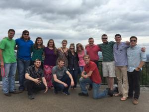 A group of thirteen people, with eight standing and five kneeling, posing together outdoors under a cloudy sky.
