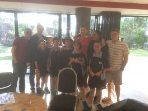A group photo with adults and children in front of large windows indoors.