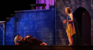 Alt-text:  Theatrical scene with one actor lying down and another standing, set against a blue stone wall backdrop.