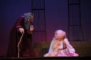 An old man with a cane stands looking at a woman on the floor with a purple background and dim lighting