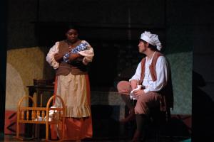 A man wearing a chefs hat sitting down talking to a woman standing up holding a newborn next to a crib in dim lighting