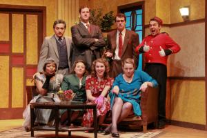 A group of eight people in vintage clothes posed on a stage set with a sofa and vintage decor.