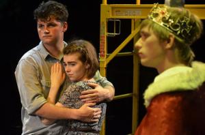 "Three individuals in a stage scene, one wearing a crown and robe, with a couple standing closely in the background."