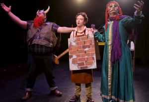 Three performers on stage in elaborate costumes, including a Viking, a brick wall, and a person in a gown.