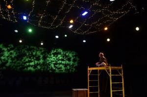 Person sitting on a scaffold on a dimly lit stage with colorful lights and string lights above them.