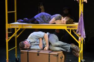 Two people sleeping on a yellow scaffolding platform and a brown box in a dark theater scene.