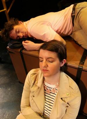 Two people resting near a suitcase, one lying on top and the other leaning against it.