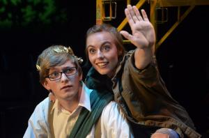 Two people on stage, one with glasses and a gold headpiece, the other raising their hand.