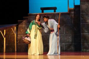 Two actors in costume on a stage, with one holding a basket and the other a staff.