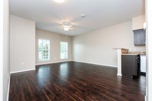Empty room with dark hardwood flooring, light beige walls, two large windows, and a kitchenette area.