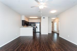 Empty apartment interior with dark wooden floors, open kitchen, and a ceiling fan.