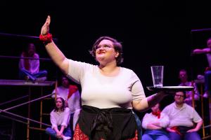 The woman holds her right hand in the air while her left holds a tray with a glass of water on top; in the background, a group of actors is sitting in Working: The Musical Theatre production.