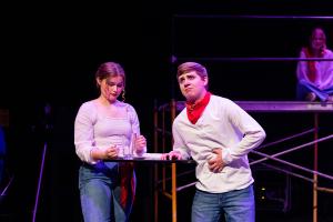 A woman on the left of a small circular table and a man to the right of the table clenching his stomach in Working: The Musical Theatre production.