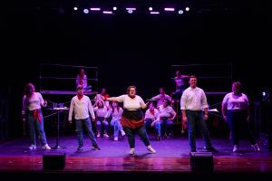 Five actors in the front of the stage behind them are a group of actors sitting down in Working: The Musical Theatre production. 