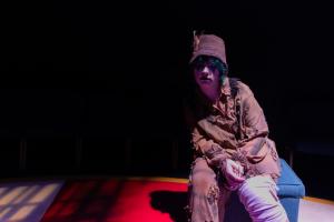 Person in a brown outfit and hat sitting on a stage with a spotlight.
