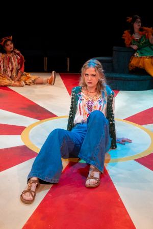  Young woman in colorful, bohemian outfit seated on a vibrant red and white circular patterned floor, with other performers in the background.