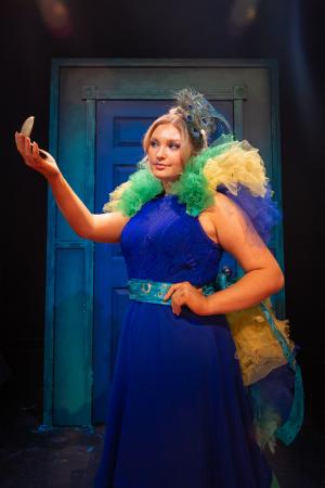 Woman looking at herself in handheld mirror in front of blue door during Cagebirds Theatre Production
