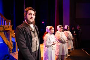 Five people in historical attire stand on a stage under dramatic lighting.
