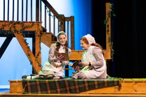  Two individuals in period costumes reading books on a stage platform with a staircase in the background