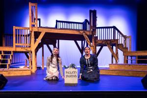 Two actresses sit on a theater stage in front of a wooden structure, with a box labeled "RUSTY BLOSSUMS" between them.