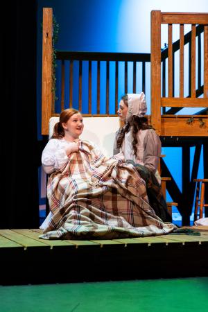 Two people sitting on a wooden porch structure during a stage performance, one with a checkered blanket.