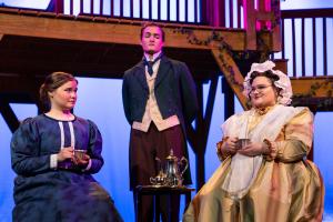Three actors in period costumes, with two seated and one standing between them, set against a stage background with stairs and railings.