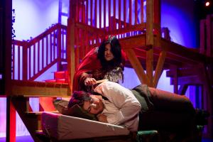  Two individuals on a stage; one person bends over another who is lying down on a bench, with dramatic red and purple lighting highlighting wooden structures around them.