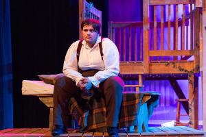 Person in white shirt and suspenders sitting on a bench on a stage with a wooden structure in the background under blue and purple lighting.