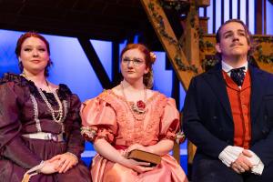 Three people in historical costumes seated in front of a wooden staircase and a blue-lit background.
