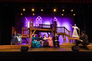 A theater stage with actors in period costumes performing, with a two-level wooden structure and purple-lit arch windows in the background.