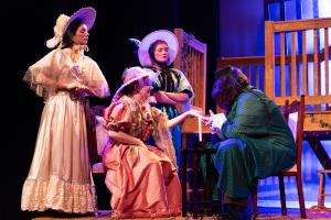 Four people in period costumes performing on stage around a table with vibrant lighting.