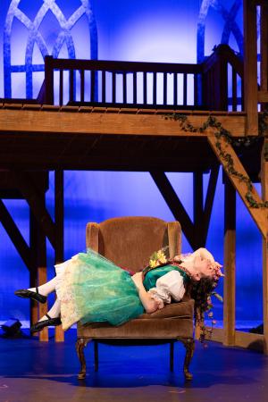 Child in a green dress reclining sideways on an armchair on a theatrical stage with a wooden balcony in the background.