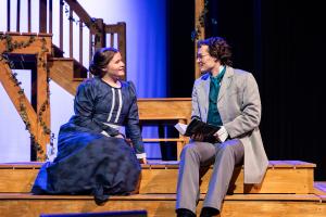 Two actors in period costumes sit on wooden steps on stage, engaged in conversation.