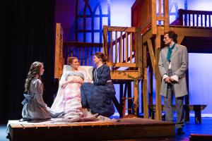  Theatrical stage with four actors in period costumes, gathered around a bed with a wooden structure in the background.