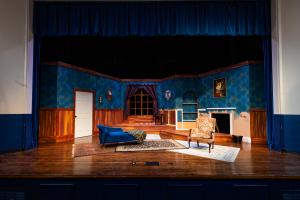 Stage set of a luxurious living room with blue walls, wood accents, sofa, armchair, and fireplace.