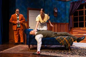 Three actors on stage; one lies on a couch, a woman leans over him, and another man stands watching.