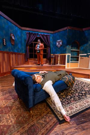 A theatrical scene with a person lying on a rug and another standing in the background holding a glass.