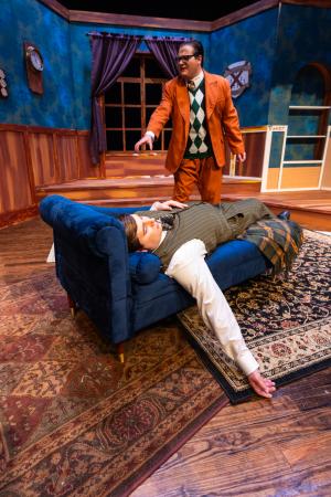 A theatrical scene with a person lying on a rug and another standing in the background holding a glass.