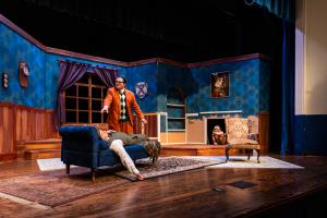 Theater stage set as a living room with blue walls and wooden accents, featuring two men, one standing and one lying on a sofa.