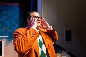 Person on stage wearing an orange blazer and green argyle sweater with an expression of surprise.