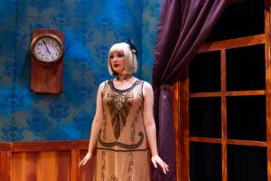 Person in a flapper dress stands in a vintage room with a wooden clock and large draped window.