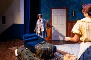 Theatrical scene featuring three actors, a chaise lounge, and a door on a stage.
