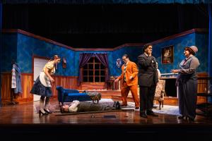 Cast of actors performing in a period-piece stage play, set in an elegantly furnished room.