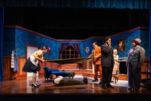 Cast of actors performing in a period-piece stage play, set in an elegantly furnished room.