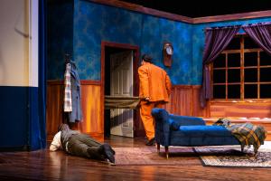 A stage set of a room with blue and orange décor, featuring two men, one lying on the floor and one standing in a doorway, with a blue chaise lounge by a window.