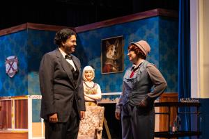 Three actors on stage, two in the foreground conversing in costumes, and one in the background.