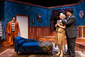 A theatrical stage set with actors in a vintage-style room featuring a chaise lounge, blue walls, and wood paneling.