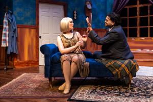 A theatrical stage set with actors in a vintage-style room featuring a chaise lounge, blue walls, and wood paneling.