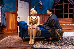 A theatrical stage set with actors in a vintage-style room featuring a chaise lounge, blue walls, and wood paneling.